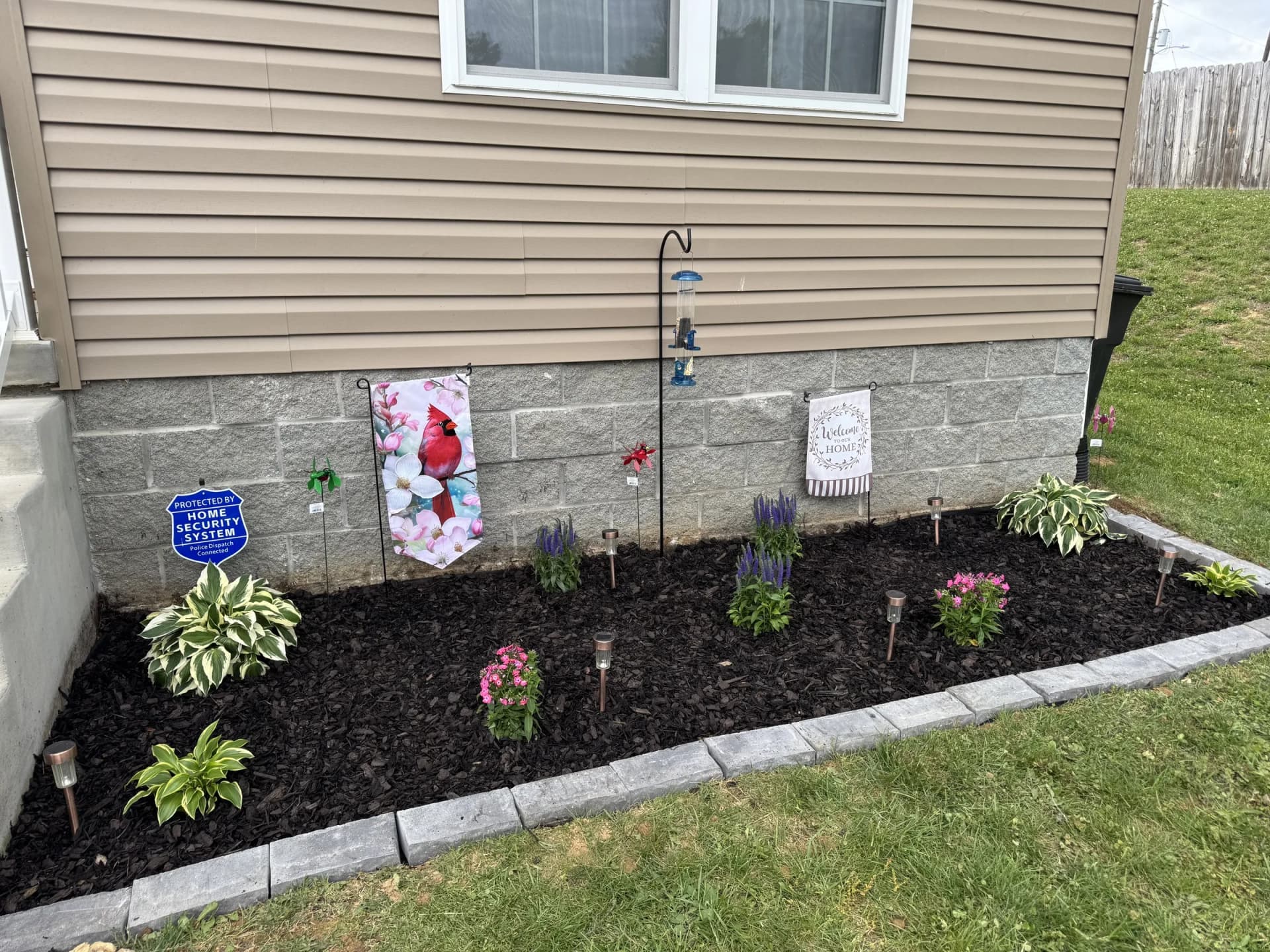 Dark mulched flower bed with hostas and colorful flowering plants, edged with gray stone blocks beside a house.